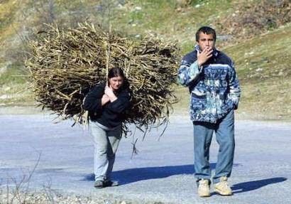 man smokes while wife walks behind carrying heavy load